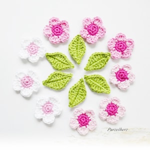 Könnte beinhalten: Eine Sammlung gehäkelter rosa und weißer Blumen und grüner Blätter. Die Blumen haben eine rosa Mitte und sind in einem Kreis um die Blätter angeordnet.