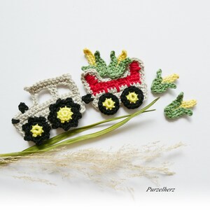 Könnte beinhalten: Häkelspielzeug-Set mit einem grauen Traktor mit schwarzen Rädern und gelben Zentren, einem roten Wagen mit grünen Maisstängeln und drei einzelnen Maisstängeln. Die Spielzeuge sind auf einem weißen Hintergrund mit einem getrockneten Grashalm angeordnet.