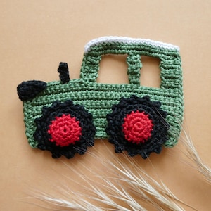 Könnte beinhalten: Eine gehäkelte Traktor-Applikation in Olivgrün mit weißem Dach, schwarzen Rädern und leuchtend roten Mitten. Der Traktor verfügt über ein kleines schwarzes Auspuffrohr auf der Motorhaube und ein ausgeschnittenes Fensterdesign aus Garn.