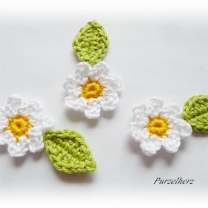 Könnte beinhalten: Gehäkelte weiße Gänseblümchenblüten mit gelben Zentren und grünen Blättern. Die handgefertigten floralen Verzierungen sind auf weißem Hintergrund angeordnet. Der Text "Purzelherz" befindet sich in der unteren rechten Ecke.