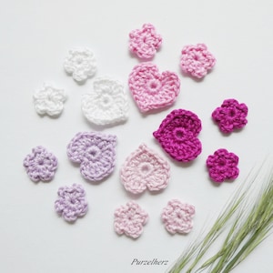 Könnte beinhalten: Eine Sammlung handgefertigter Häkelblumen und -herzen in verschiedenen Rosa-, Lila- und Weißtönen. Das Bild zeigt eine Vielzahl von Blumen- und herzförmigen Designs, ideal für Bastelprojekte. Das Purzelherz-Logo ist unten sichtbar.