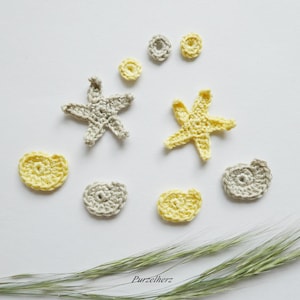 Könnte beinhalten: Handgefertigte Häkelarbeiten in Gelb- und Grautönen, darunter Seesterne, Muscheln und kreisförmige Formen, angeordnet auf einer weißen Oberfläche. Einige grüne Grashalme sind zu sehen. Das Wort "Purzelherz" steht unten.