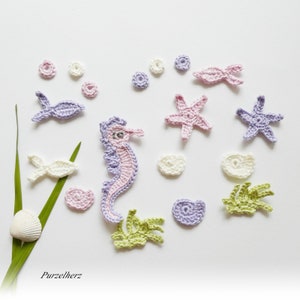 Könnte beinhalten: Gehäkelte Meerestiere in Pastellfarben, darunter ein Seepferdchen, Seesterne, Fische und Muscheln. Das Seepferdchen ist lavendelfarben und rosa, die Seesterne sind rosa und lavendelfarben. Grünes Seegras und weiße Blasen sind ebenfalls vorhanden.