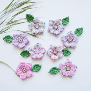 Könnte beinhalten: Eine Sammlung handgefertigter Häkelblumen in verschiedenen Pastellfarben, darunter Rosa, Lila und Blau, jede mit einem grünen Blatt. Die Blumen sind auf einer weißen Oberfläche angeordnet und zeigen ihre komplizierten Details und ihre filigrane Handwerkskunst.