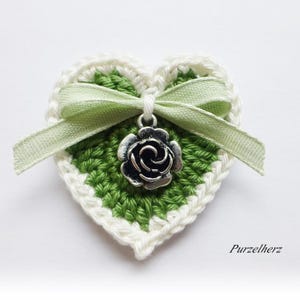 Könnte beinhalten: Ein grünes Häkelherz mit weißem Rand und einem grünen Satinband. Ein silberner Anhänger in Form einer Rose hängt in der Mitte des Herzens.
