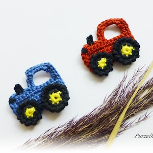 Könnte beinhalten: Zwei gehäkelte Traktor-Ornamente, eines in Blau und eines in Rot, mit schwarzen und gelben Rädern. Die Traktoren sind vor einem weißen Hintergrund mit getrocknetem Gras und dem Text "Purzelherz" platziert.