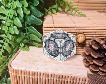 May include: A white, gray, and pink cube-shaped sticker with a geometric pattern and two pink hearts. The sticker is on a wooden surface.