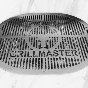 Può includere: Un'immagine in bianco e nero di una griglia di barbecue rotonda in metallo con il testo "Barbad's Grillmaster" in rilievo.