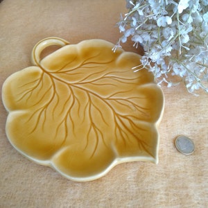 May include: A ceramic leaf-shaped dish in a warm yellow colour. The dish features detailed vein patterns and a small handle. The design is reminiscent of nature, perfect for serving or display. The dish is approximately 20 cm long.