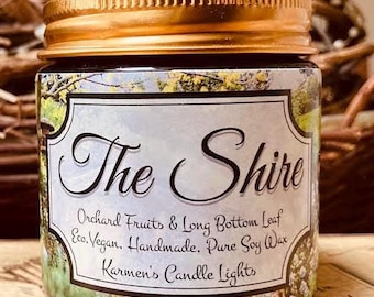 May include: A glass candle jar with a gold-colored metal lid. The label features a green landscape illustration and the text: The Shire, Orchard Fruits & Long Bottom Leaf, Eco, Vegan, Handmade, Pure Soy Wax, Karmen's Candle Lights.