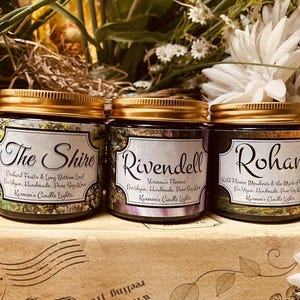 May include: Three cylindrical glass candle jars with gold-coloured screw-top lids. Each jar features a white label with black text reading The Shire, Rivendell, and Rohan, along with additional descriptive text and the brand name Karmen's Candle Lights.