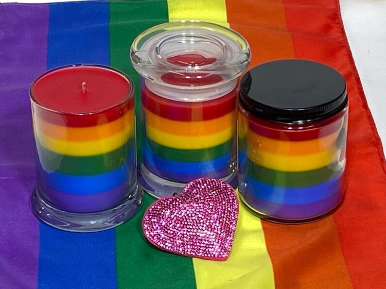 Op de afbeelding: Drie regenboogkleurige gelaagde kaarsen in glazen potten met deksels. De kaarsen zijn rood, oranje, geel, groen, blauw en paars. Een roze hartvormig decoratief object bevindt zich voor de kaarsen.