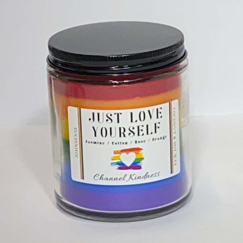 Op de afbeelding: Een glazen kaars pot met een zwarte deksel. De kaars is gelaagd met de kleuren van de regenboog en heeft de tekst "JUST LOVE YOURSELF" met de woorden "Jasmine / Cotton / Rose / Orange" en "Channel Kindness" eronder. De kaars is gemaakt van kokosnoot en sojawas.