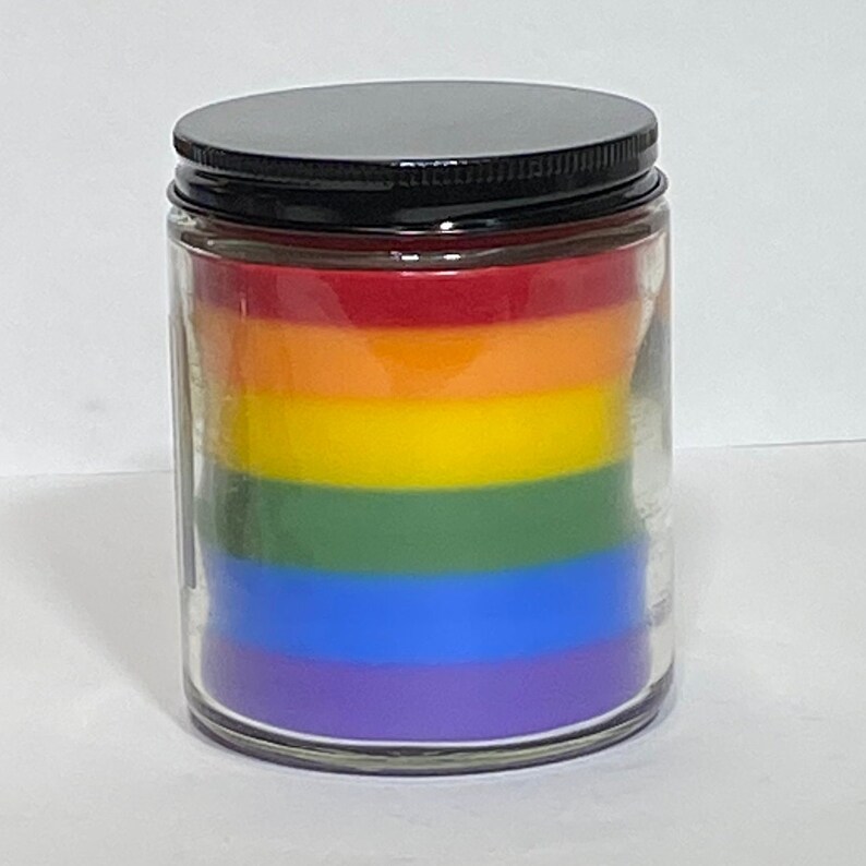 Op de afbeelding: Een regenboogkleurige gelaagde kaars in een glazen pot met een zwarte deksel. De kaars is gelaagd met rode, oranje, gele, groene, blauwe en paarse was.