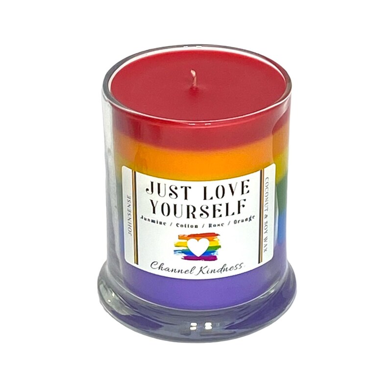 Op de afbeelding: Een regenboogkleurige gelaagde kaars in een glazen pot met de tekst "JUST LOVE YOURSELF" en "Jasmine / Cotton / Rose / Orange" en "Channel Kindness".