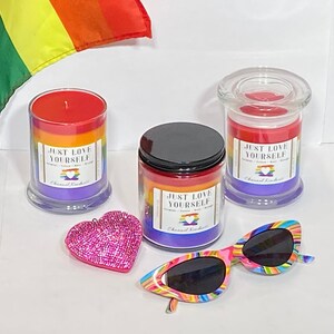 Op de afbeelding: Drie regenboogkleurige kaarsen met de tekst "Just Love Yourself" op de etiketten. De kaarsen staan in glazen potten met een regenboogontwerp. Er is een roze, glinsterende hartvormige versiering en een paar regenboogkleurige zonnebrillen op de foto.