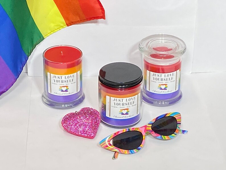 Op de afbeelding: Drie regenboogkleurige kaarsen met de tekst "Just Love Yourself" op de etiketten. De kaarsen staan in glazen potten met een regenboogontwerp. Er is een roze, glinsterende hartvormige versiering en een paar regenboogkleurige zonnebrillen op de foto.