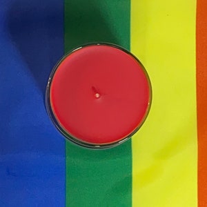 Op de afbeelding: Een rode kaars in een glazen pot, op een regenboogkleurige stoffen achtergrond.