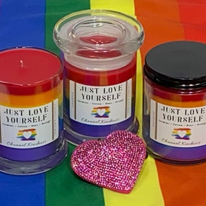 Op de afbeelding: Drie glazen kaarsenpotten met regenboogkleurig was en de tekst "Just Love Yourself" op het etiket. De kaarsen staan op een regenboogkleurige stoffen achtergrond.