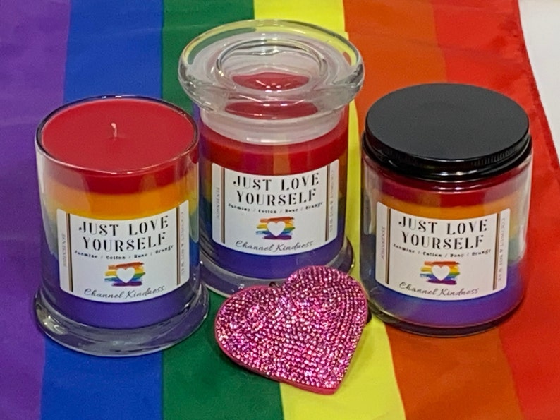Op de afbeelding: Drie glazen kaarsenpotten met regenboogkleurig was en de tekst "Just Love Yourself" op het etiket. De kaarsen staan op een regenboogkleurige stoffen achtergrond.