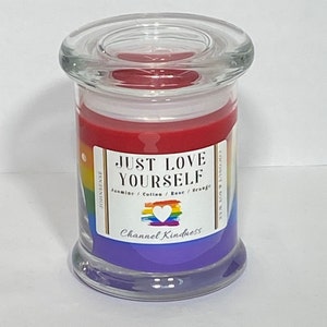 Op de afbeelding: Een glazen pot met een regenboog-pride-vlagontwerp en de tekst "Just Love Yourself". De kaars is een mengsel van kokos- en sojawas met een geur van jasmijn, katoen, roos en sinaasappel. Het etiket zegt ook "Channel Kindness".