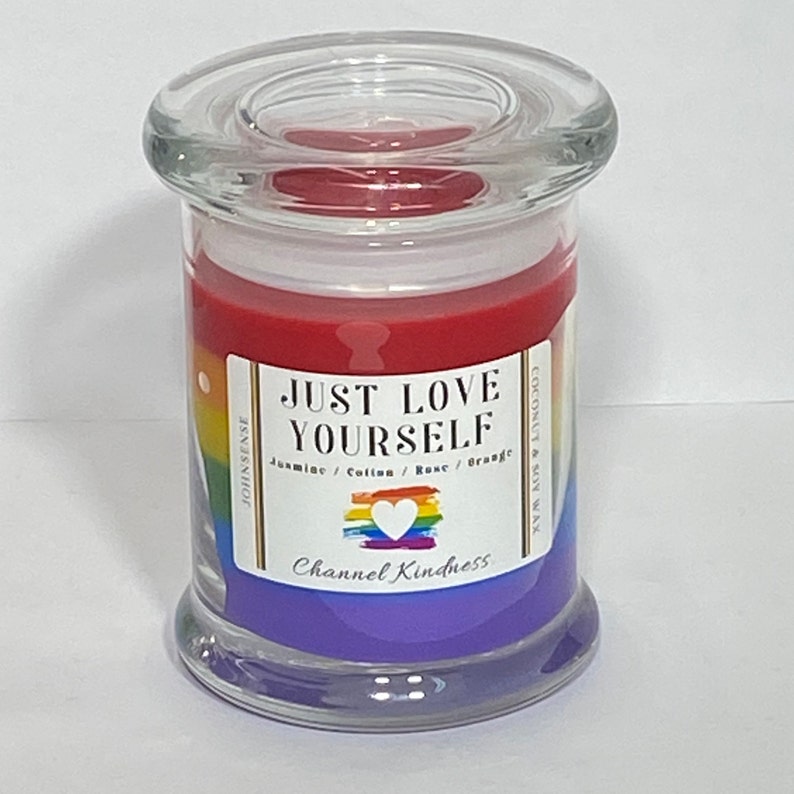 Op de afbeelding: Een glazen pot met een regenboog-pride-vlagontwerp en de tekst "Just Love Yourself". De kaars is een mengsel van kokos- en sojawas met een geur van jasmijn, katoen, roos en sinaasappel. Het etiket zegt ook "Channel Kindness".