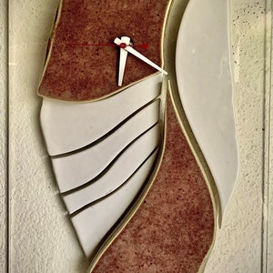 May include: A red and white ceramic clock with a unique abstract design. The clock face is made of white ceramic with a red hour hand and a white minute hand. The clock is mounted on a white wall.