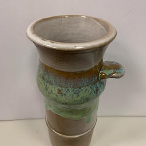 May include: A handmade ceramic vase with a unique shape and a small handle. The vase features a color palette of brown, green, and blue, with a white rim. The vase is approximately 7 inches tall.