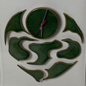 May include: A green ceramic clock with a white face and black hands. The clock is surrounded by a design of green ceramic shapes with gold accents. The clock is signed "P. Ball" in the bottom right corner.