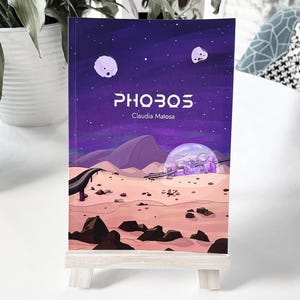 May include: A book cover with a purple background and a city inside a bubble on a desert planet. The title of the book is "PHOBOS" by Claudia Matosa.