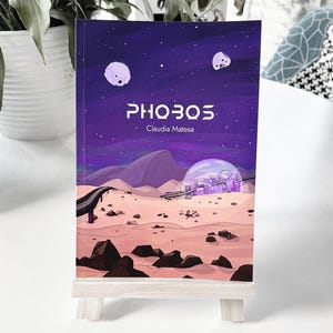 May include: A purple book cover with the title "PHOBOS" and the author's name "Claudia Matosa". The cover features a cartoon illustration of a city inside a bubble on a desert planet with a purple sky and two white moons.