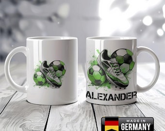 Könnte beinhalten: Weiße Keramik-Tasse mit Fußball-Motiv. Das Design zeigt einen Fußballschuh, zwei Fußbälle und grüne Farbspritzer. Der Name "ALEXANDER" ist auf der Tasse aufgedruckt. Ein "Made in Germany"-Aufkleber ist sichtbar.