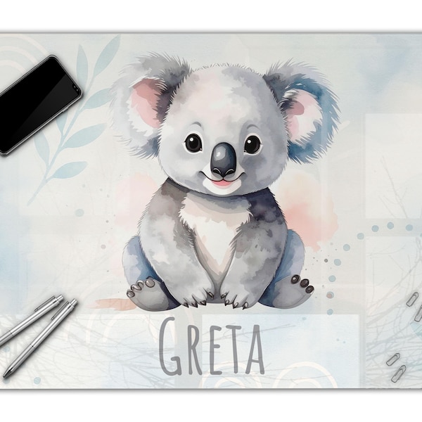 Pode incluir: Um tapete de secretária retangular com uma ilustração em aguarela de um coala cinzento centrado num fundo azul claro e branco. O nome GRETA está impresso em letras maiúsculas cinzentas por baixo do coala. Um smartphone preto e duas canetas prateadas estão colocados sobre o tapete.