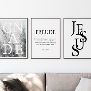 Könnte beinhalten: Drei gerahmte Kunstdrucke mit schwarzen Rahmen. Der linke Druck zeigt das Wort "GNADE" in großen weißen Buchstaben über einem Graustufenbild von hohem Gras. Der mittlere Druck zeigt "FREUDE" mit deutschem Text darunter. Der rechte Druck zeigt "JESUS" in großen schwarzen Buchstaben.