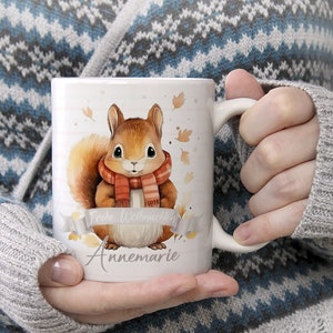 Könnte beinhalten: Weiße Keramiktasse mit einem braunen Eichhörnchen, das einen rot-weißen Schal trägt. Auf der Tasse steht "Frohe Weihnachten Annemarie" gedruckt.