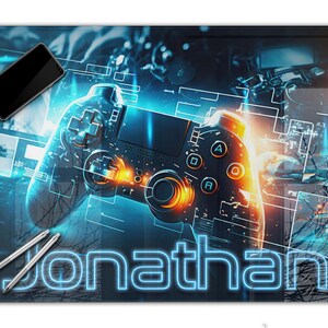 Könnte beinhalten: Eine rechteckige Schreibtischunterlage mit einem zentralen schwarzen Videospiel-Controller mit leuchtenden blauen und orangefarbenen Lichteffekten, einem Smartphone in der oberen linken Ecke, zwei silbernen Stiften und dem Namen Jonathan in großen, hellblauen Neonbuchstaben.