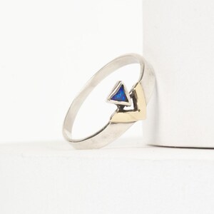Könnte beinhalten: Ein silberner Ring mit goldenem Akzent und einem blauen, dreieckigen Stein. Der Ring ist vor einem weißen Hintergrund platziert, was sein modernes Design und seine geometrische Form hervorhebt. Das Band des Rings hat eine strukturierte Oberfläche.