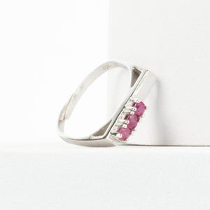 Könnte beinhalten: Ein silberfarbener Metallring mit einer Reihe von drei kleinen, runden, leuchtend rosaroten Edelsteinen, die vertikal entlang der Ringschiene gefasst sind. Der Ring hat eine polierte Oberfläche und eine schmale Schiene, die nach hinten zuläuft.