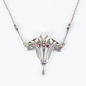 Op de afbeelding: Zilverkleurige ketting met een Art Nouveau-hanger. De hanger heeft een bloemmotief met twee rode edelstenen en een druppel. De ketting heeft een fijne ketting en staat tegen een witte achtergrond.