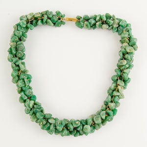 Op de afbeelding: Een ketting bestaande uit kleine, onregelmatig gevormde groene stenen. De stenen zijn geregen met goudkleurig draad en kleine goudkleurige accenten. De ketting wordt getoond tegen een witte achtergrond.