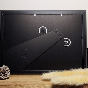 May include: Black picture frame with a black easel stand on a wooden surface. The frame has a white paper backing and a white mat.