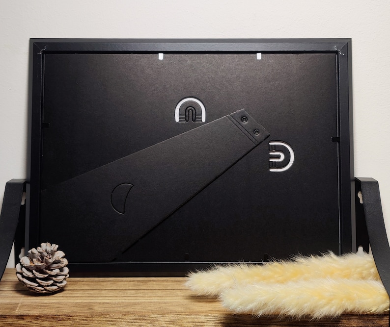 May include: Black picture frame with a black easel stand on a wooden surface. The frame has a white paper backing and a white mat.