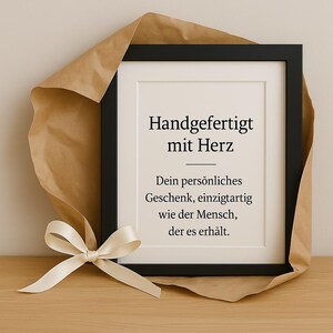 May include: A framed print with the text "Handgefertigt mit Herz" (Handmade with heart) and a message about a personalized gift. The print is sitting on a brown paper surface with a white ribbon.