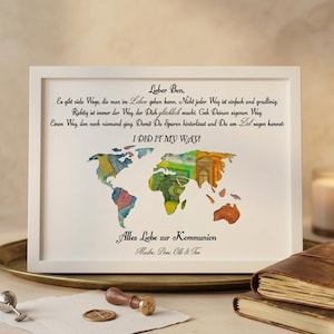 Könnte beinhalten: Ein gerahmter Druck mit einer Aquarell-Weltkarte, die farbenfrohe Währungen enthält. Der Text "I DID IT MY WAY!" ist zu sehen, zusammen mit deutschem Text. Auf einem Tisch mit Kerze und ledergebundenem Buch.