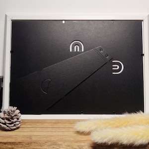 May include: White picture frame with a black mat and a white easel stand on a wooden surface. The frame is sitting on a wooden table with a pine cone and dried grass.
