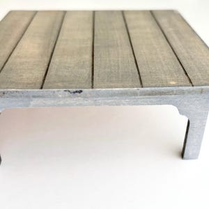 May include: A small, rectangular wooden table with a gray finish. The tabletop has a slatted design, and the legs are a matching gray color. The table is approximately 12 inches long and 8 inches wide.