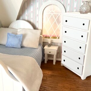 May include: A white bed with a blue and white checked pillow and a white blanket. A white bedside table with a lamp is next to the bed. A white chest of drawers with black knobs is in the corner of the room. The room has striped wallpaper with pink flowers.