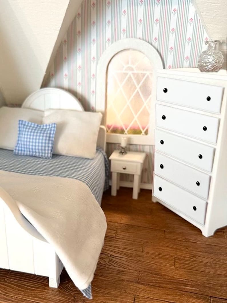 May include: A white bed with a blue and white checked pillow and a white blanket. A white bedside table with a lamp is next to the bed. A white chest of drawers with black knobs is in the corner of the room. The room has striped wallpaper with pink flowers.
