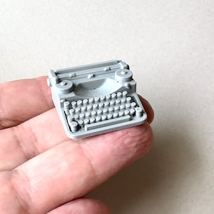 May include: A small, gray, plastic model of a vintage typewriter. The typewriter has a round, black, plastic roller and a keyboard with round, black, plastic keys.