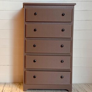 May include: A brown wooden chest of drawers with five drawers and black knobs. The chest of drawers is standing on a light wood floor.